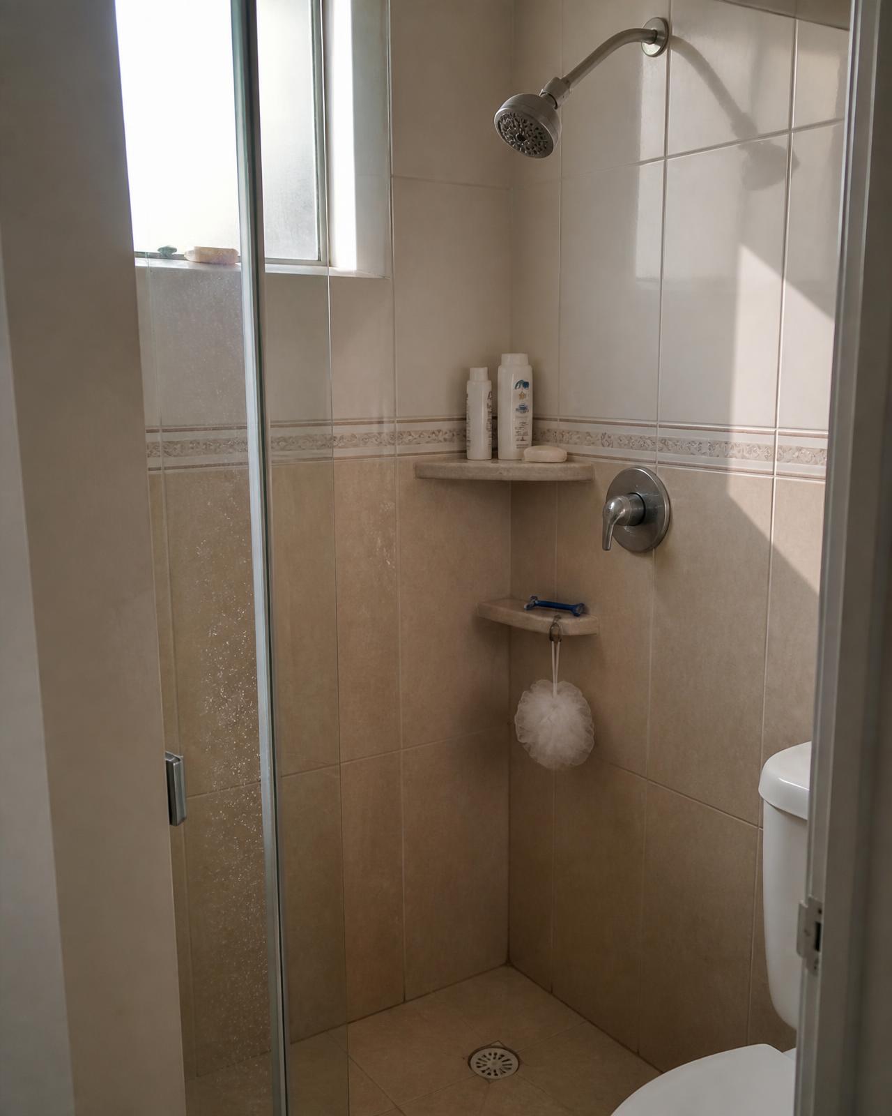 Compact tiled shower in a tiny apartment with shampoo, soap and a razor on a small corner shelf